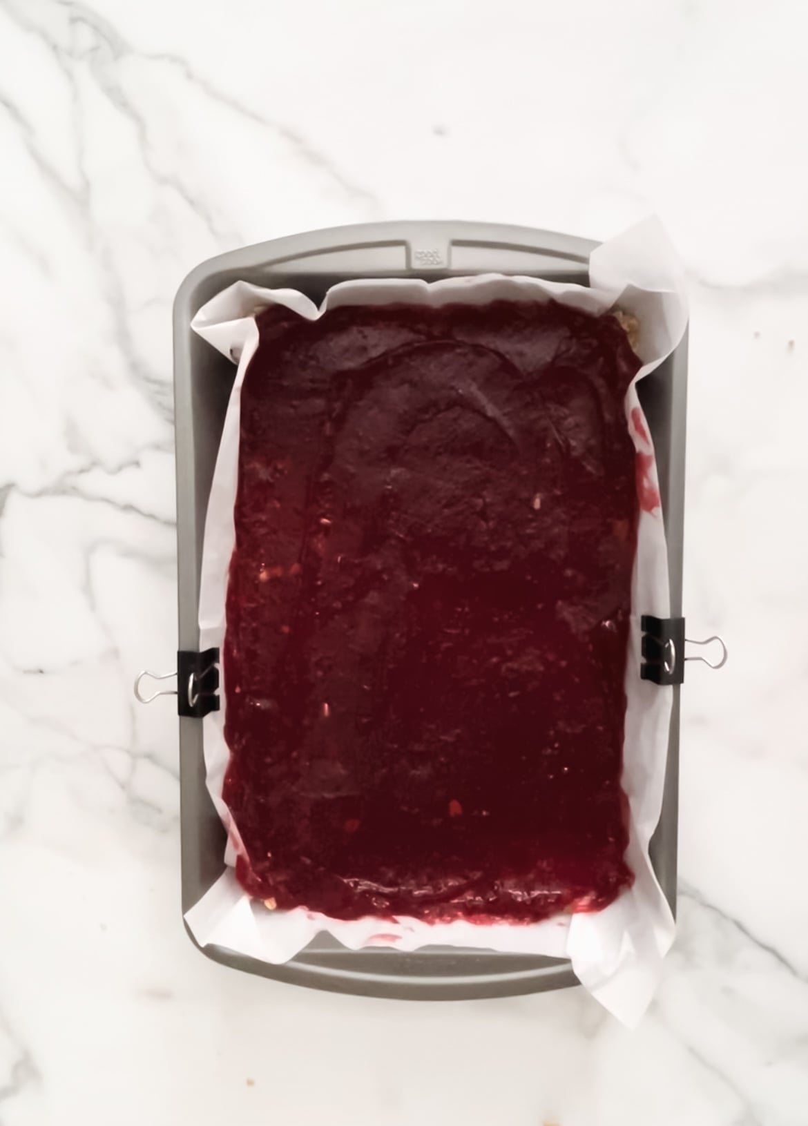The base of the raspberry oatmeal bars in a baking pan, topped with raspberry jam.